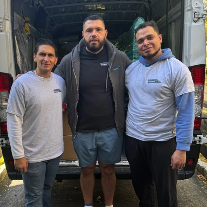 Moving company owner Joseph Camaj of Camaj & Go movers with two crew members. All three people are standing in front of a moving van in Westchester wearing Camaj & Go movers tshirts.